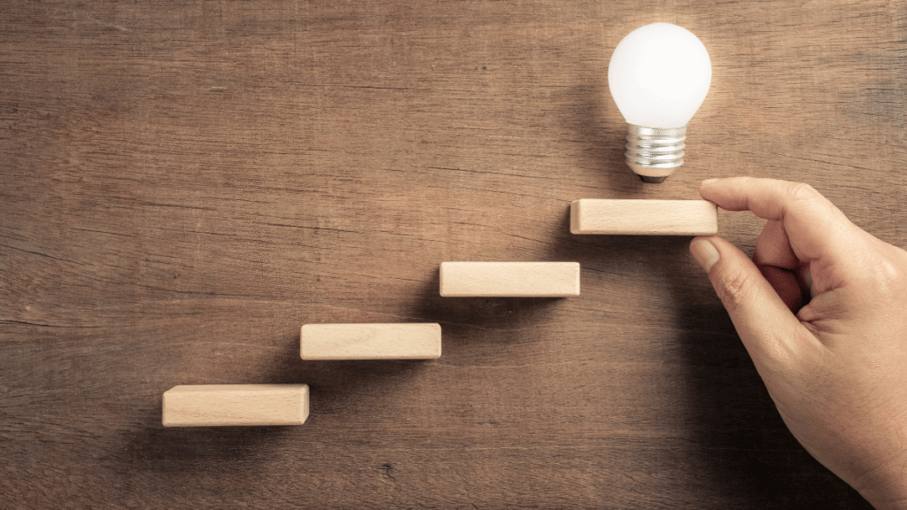 Dłoń układająca stopnie z klocków prowadzące do żarówki – symbol małych kroków do celu