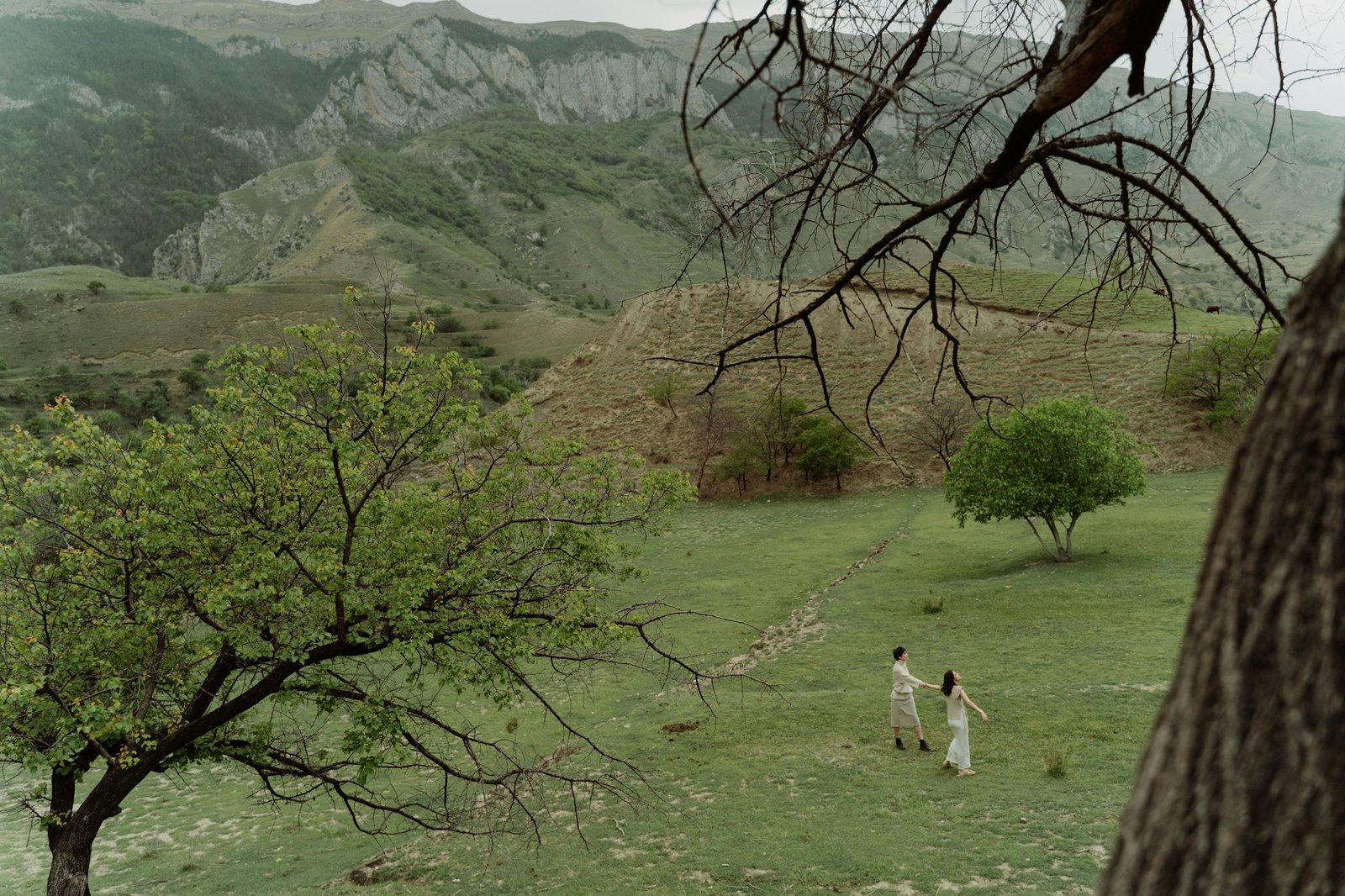 A couple walks hand in hand through a lush green valley surrounded by picturesque mountains.